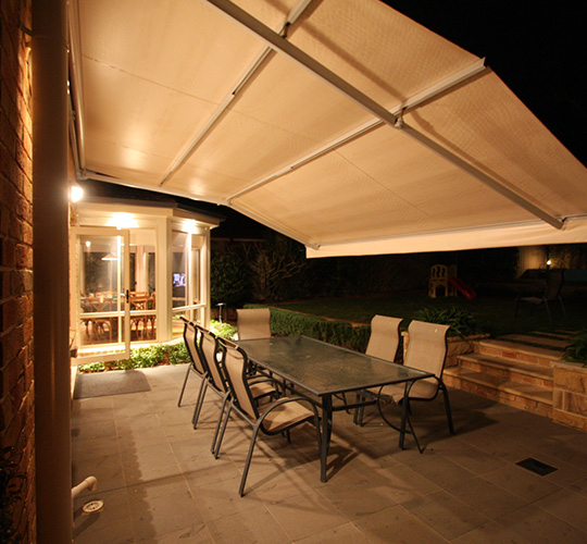 Nächtlich beleuchtete Terrassse mit ausgefahrener Markise über einer Essgruppe