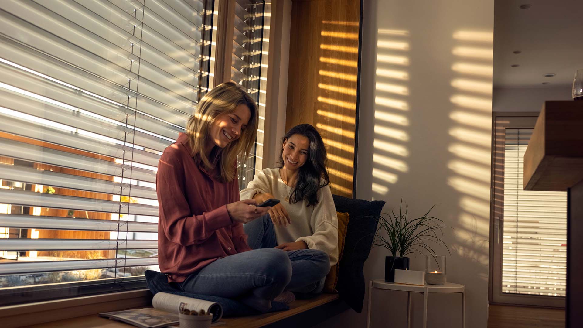 Zwei Frauen sitzend auf einer Fensterbank mit halbgeschlossenen Jalousien im Hintergrund