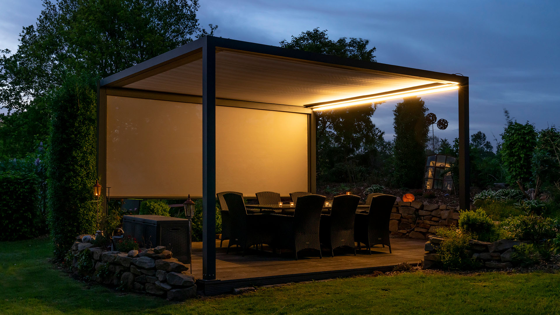 Terrassenmobiliar Essgruppe unter Lamellenüberdachung Sitzgruppe unter einer Textilterassenüberdachung bei abendlicher stimmungsvoller Beleuchtung